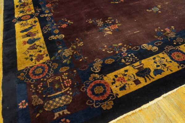 A low-pile burgundy rug with patterned gold, navy, and multi-colored borders rests on yellow wood flooring.