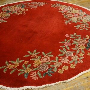 Circular red rug with a floral garland pattern and white fringe on distressed wood planks.