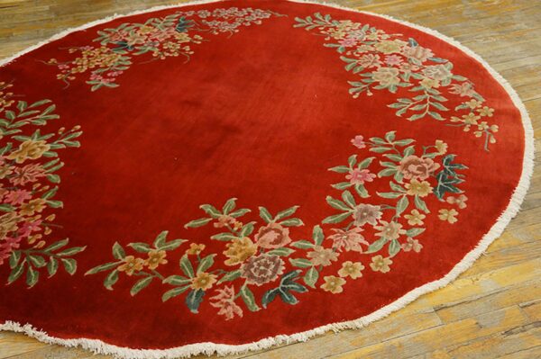 Circular red rug with a floral garland pattern and white fringe on distressed wood planks.