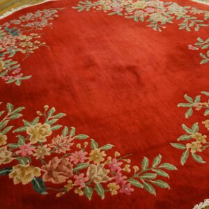 Circular deep red rug with high pile and multicolored floral garland rests on wood flooring.