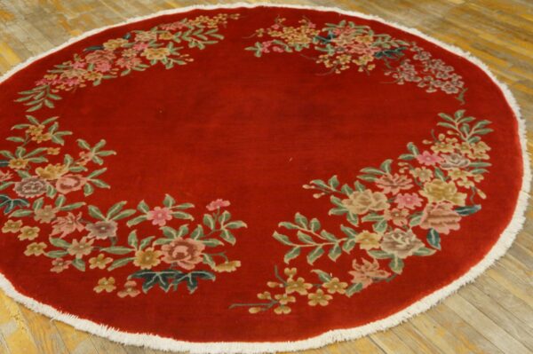 Circular deep red rug with colorful floral sprays around the edge, white fringe, resting on wood planks.