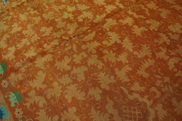 Rust and gold low-pile rug with repeating large-scale botanical designs; a green border section is visible.