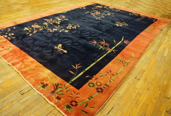 Navy blue rug with floral and bamboo motifs, a wide orange border, and visible sheen on yellow wood flooring.