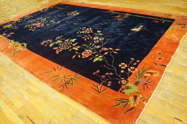 Plush navy rug with floral pattern and wide orange border lies on reflective light wood floors.
