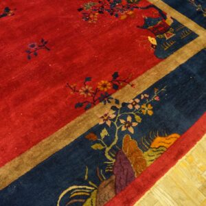 Red field rug with blue border featuring floral and pagoda patterns rests on light wood planks.