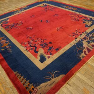 Ornate red rug featuring blue landscape borders and tan geometric framing on worn light brown wood planks.