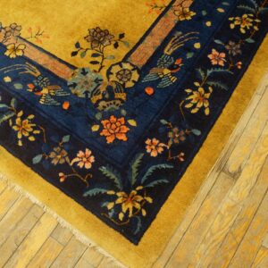 Detail of a gold rug border with dark blue floral and bird motifs on light wood flooring.