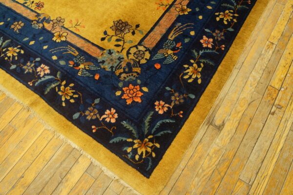 Detail of a gold rug border with dark blue floral and bird motifs on light wood flooring.