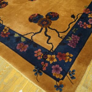Gold rug with a navy border displays large floral patterns in rust, purple, and blue on wood planks.