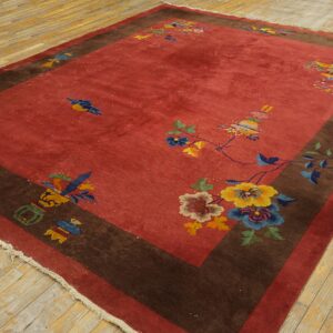 A red field rug with a wide brown border and sparse multi-colored floral designs lies on light wood planks.
