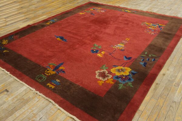 A red field rug with a wide brown border and sparse multi-colored floral designs lies on light wood planks.
