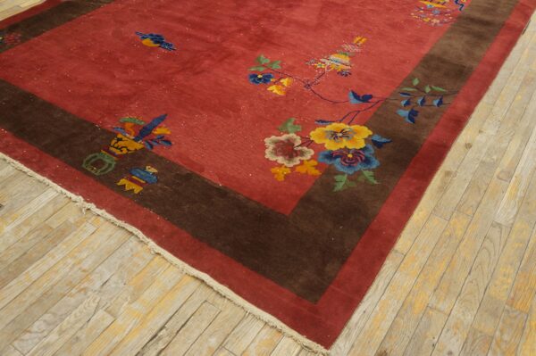 Red rug with stylized multi-colored florals and brown border rests on distressed wood flooring.