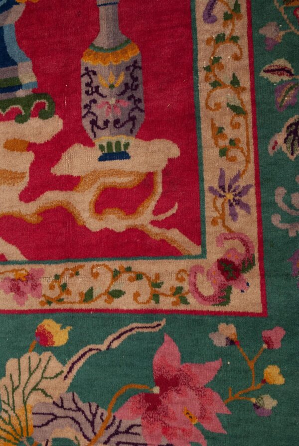 Close-up of an ornate rug featuring a magenta field, a gray vase motif, and a wide floral border on teal green.