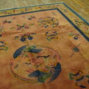 Salmon rug with colorful mythological patterns, contrasting blue border, set on distressed wood flooring.