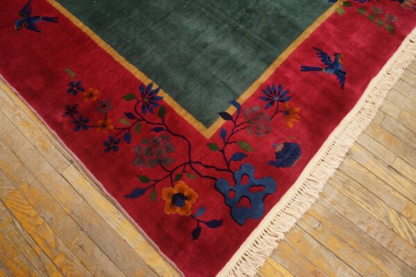 High-pile rug corner showing a green field, red floral border, cream fringe, and distressed wood floor.