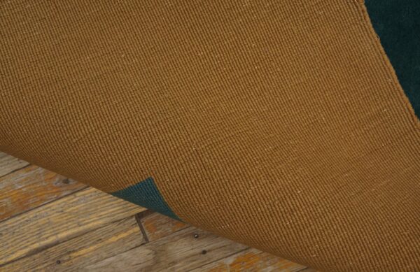 The ribbed brown rug backing is lifted, showing a dark green layer and distressed wood planks below.