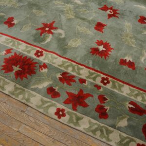 Diagonal view of a sage green plush rug with large red floral motifs resting on worn wood planks.