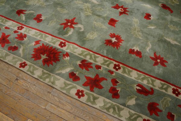 Diagonal view of a sage green plush rug with large red floral motifs resting on worn wood planks.