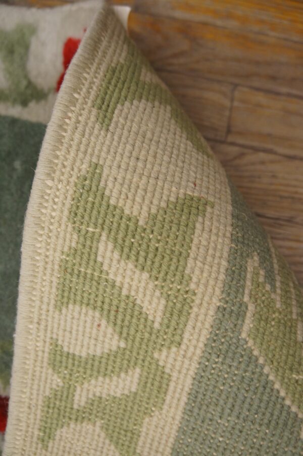 A rolled rug shows its tight beige and sage woven back, with glimpses of red pile above wood flooring.