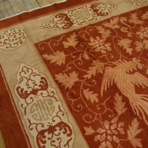 Terracotta and tan rug with a phoenix and leaf pattern borders worn wooden floorboards.