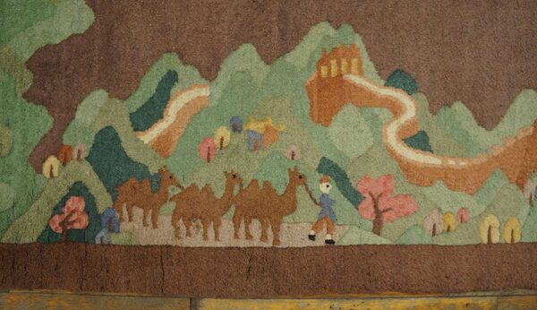 Deep pile rug showing a brown and green landscape with camels and an orange architectural structure over wood floor.