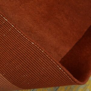 A rust-colored low-pile rug is curled, exposing the ribbed edge binding on worn yellow wood flooring.