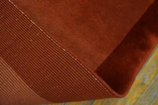 A rust-colored low-pile rug is curled, exposing the ribbed edge binding on worn yellow wood flooring.