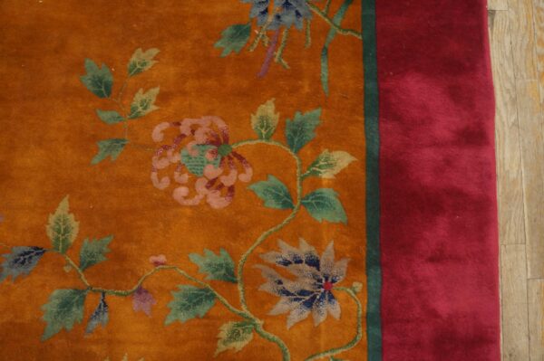 Burnished orange rug detail shows floral vines and a wide, solid red border next to wood flooring.