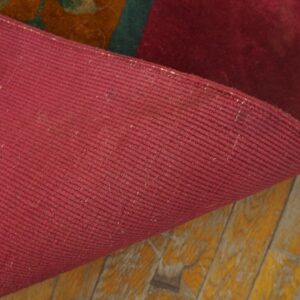 A deep red rug is curled back, exposing its tightly woven backing above distressed yellow hardwood flooring.
