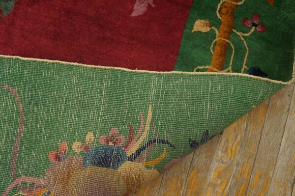 A flipped rug corner reveals a woven green backing and dense pile pattern in red, green, and brown floral elements on wood planks.