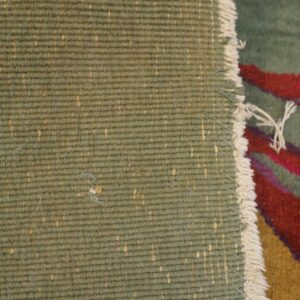 Rug detail showing the woven green backing with tan flecks, white edge fringe, and multicolor pile.