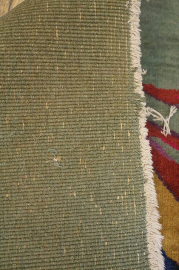 Rug detail showing the woven green backing with tan flecks, white edge fringe, and multicolor pile.
