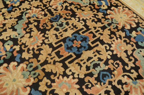 Black rug with large tan scrolling floral motifs, blue accents, and geometric fretwork on wood floor.