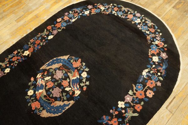 An oval black rug features a colorful floral and bird medallion and curved border on light wood flooring.