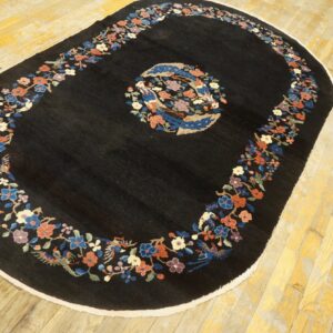 Black oval rug with colorful floral and avian borders and center medallion lying on worn yellow wood planks.
