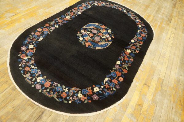 Black oval rug with colorful floral and avian borders and center medallion lying on worn yellow wood planks.