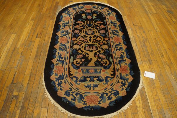 Black oval rug featuring a golden tree and vase motif bordered by coral and blue flowers on wood flooring.