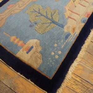Light blue scenic rug featuring foliage and structures, trimmed by a navy border on aged wood flooring.