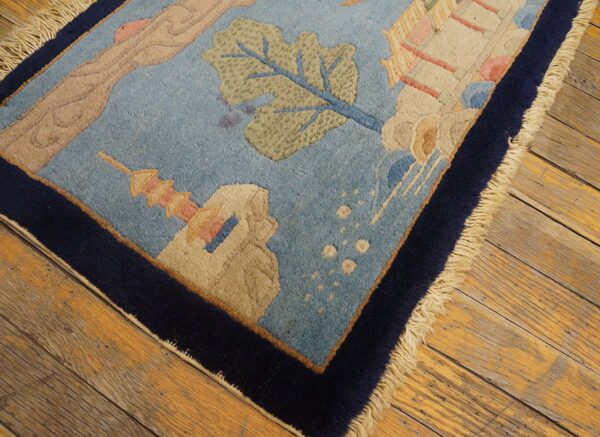 Light blue scenic rug featuring foliage and structures, trimmed by a navy border on aged wood flooring.
