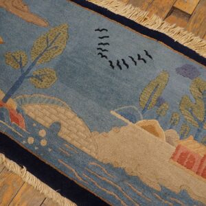 Low pile rug in blue and tan with a detailed landscape pattern, dark border, and fringe resting on rustic wood floorboards.