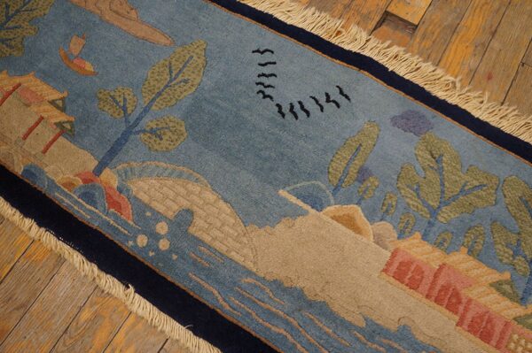 Low pile rug in blue and tan with a detailed landscape pattern, dark border, and fringe resting on rustic wood floorboards.