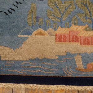 Pictorial rug corner showing blue water, tan land, pink buildings, green trees, and a navy border on a wood floor.