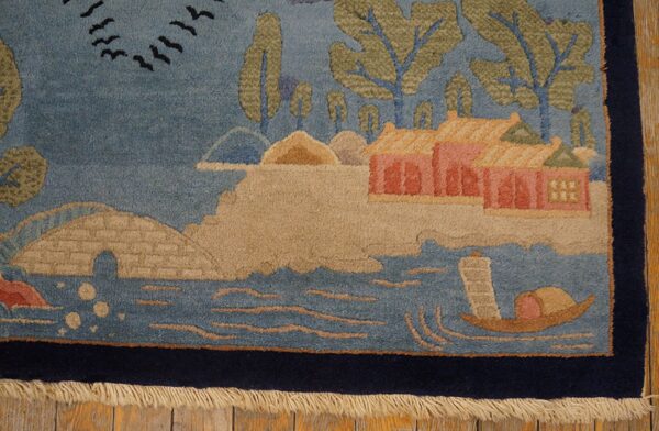 Pictorial rug corner showing blue water, tan land, pink buildings, green trees, and a navy border on a wood floor.