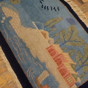 Low-pile blue rug with sculpted tan, green, and pink scenic elements, bordered in navy on distressed wood.
