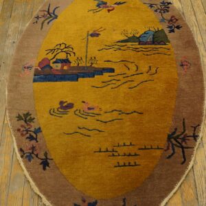 An oval mustard yellow rug with a scenic pattern and brown floral border rests on aged wood planks.