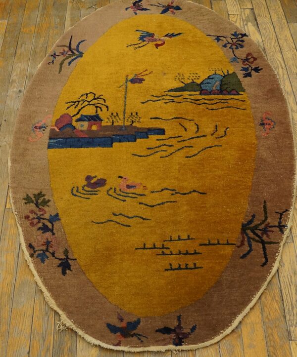 An oval mustard yellow rug with a scenic pattern and brown floral border rests on aged wood planks.