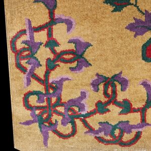 Rug close-up showing gold pile with scrolling floral motifs in purple, teal, and red.