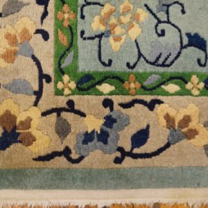 Corner view of a plush, multi-bordered rug featuring blue, tan, yellow, and green floral motifs over hardwood.