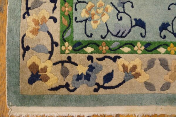 Corner view of a plush, multi-bordered rug featuring blue, tan, yellow, and green floral motifs over hardwood.