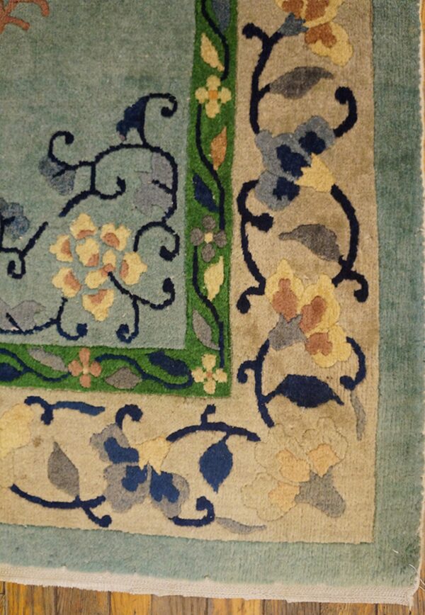Teal and beige rug corner featuring sculpted floral and vine motifs with green accents on wood floor.